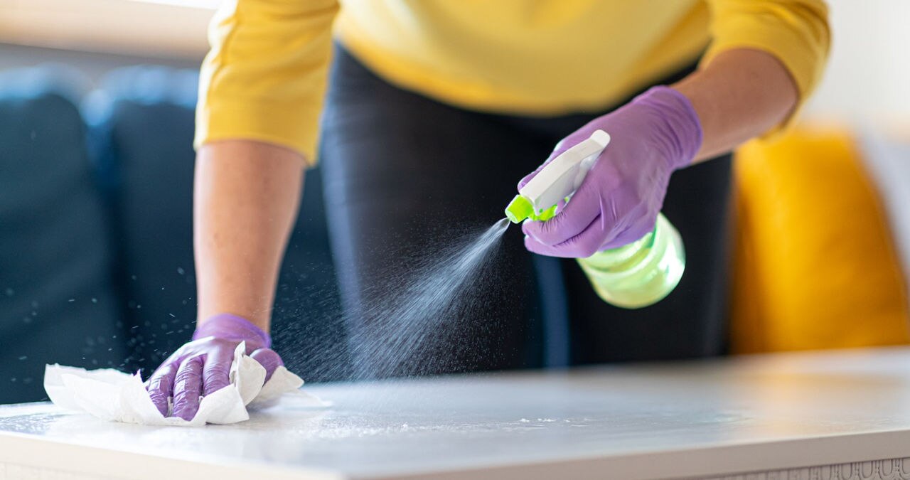 Hands in protective gloves holding spray bottle and disinfecting coffee table to wipe with paper towel against viruses